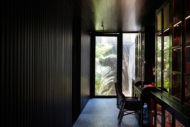 A dark, narrow interior workspace or study with black vertically ribbed wall panelling, a dark timber-planked ceiling, and a full-height glazed door or window at the end opening toward lush foliage. A compact desk and chair sit beside tall glass-fronted built-in cabinetry or shelving, with warm integrated lighting and deep green-black finishes creating a moody, intimate atmosphere.