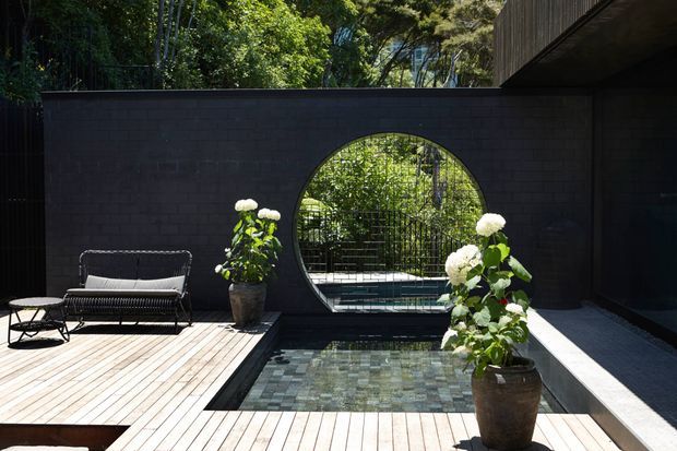 A secluded contemporary courtyard with a shallow rectangular reflecting pool set into a pale timber deck. A dark brick privacy wall features a large circular opening framing lush planting and a garden beyond. Black outdoor lounge furniture and potted white-flowering plants add contrast, while a dark-clad building edge and shaded covered area appear at right.