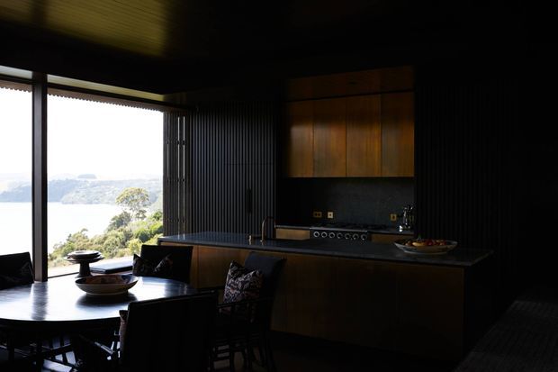 A dark, moody open-plan kitchen and dining area with a large island, integrated cooktop and sink, dark vertically grooved wall finish, and warm timber overhead cabinetry. A dining table occupies the foreground, while floor-to-ceiling glazing opens to a bright water and hillside outlook.