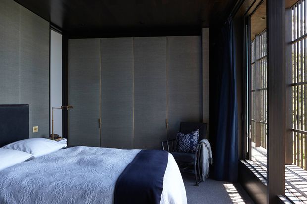 A dark, refined bedroom with a large bed in the foreground, floor-to-ceiling grey built-in wardrobe cabinetry, a deep navy ceiling and curtains, and extensive floor-to-ceiling glazing screened by vertical timber battens. The room combines strong natural light with a moody navy, grey, and white palette.