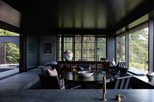 A dark, sophisticated dining and entertaining space with a round dining table, black chairs, extensive floor-to-ceiling glazing, vertical metal security or privacy screens, dark timber ceiling, and a long built-in or freestanding timber bookcase. A dark stone-topped counter with brass tapware occupies the foreground, suggesting an adjoining kitchen or bar area. The room opens toward a landscaped exterior courtyard and wooded outlook.