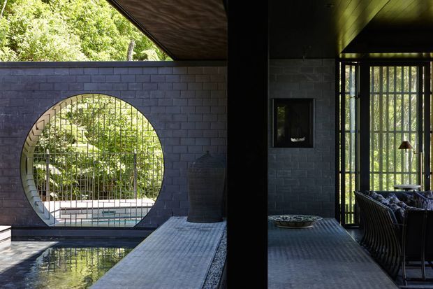 A dark, refined indoor-outdoor living space centred on a circular opening in a charcoal brick wall, with a metal security screen, lush garden beyond, and a reflecting pool in the foreground. The covered area includes a tiled floor, black structural column, vertical glazed or screened opening, built-in fireplace, and dark lounge seating. Strong geometric forms, filtered daylight, and water create a serene courtyard atmosphere.