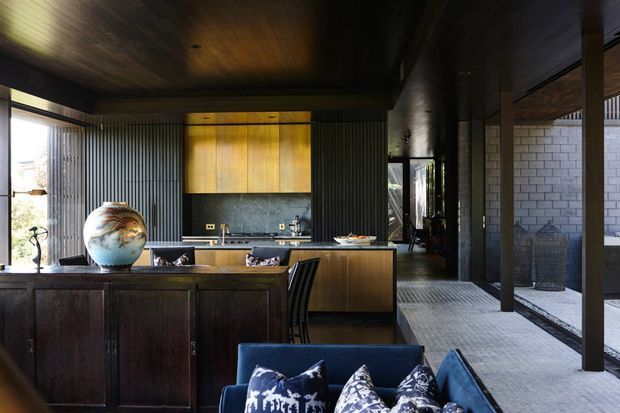 A dark, sophisticated open-plan kitchen and living area. The kitchen features a timber island with dark cabinetry, a black stone-look splashback, integrated sink and cooktop, and warm timber overhead cabinetry. Corrugated metal wall panels, a dark timber ceiling, black structural columns, and extensive glazing create an industrial-modern interior. A blue sofa and patterned cushions occupy the foreground, while a covered outdoor courtyard with pale masonry walls is visible beyond the glazing.