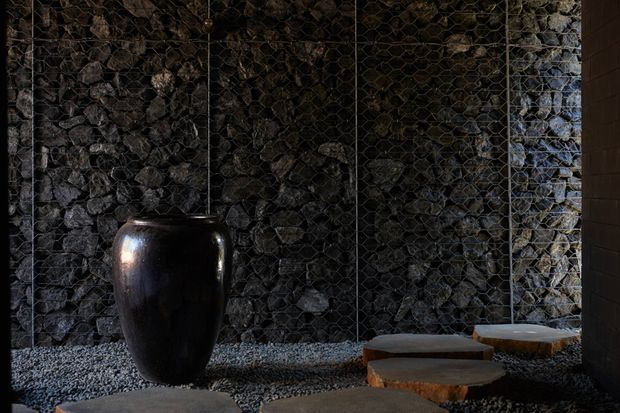 A dark, atmospheric interior or sheltered courtyard featuring a dramatic wall of irregular dark stone enclosed behind a visible wire-mesh structure. A glossy black ceramic vessel and low irregular timber stepping-stone forms sit on a bed of small grey gravel, with warm low lighting and strong natural textures.