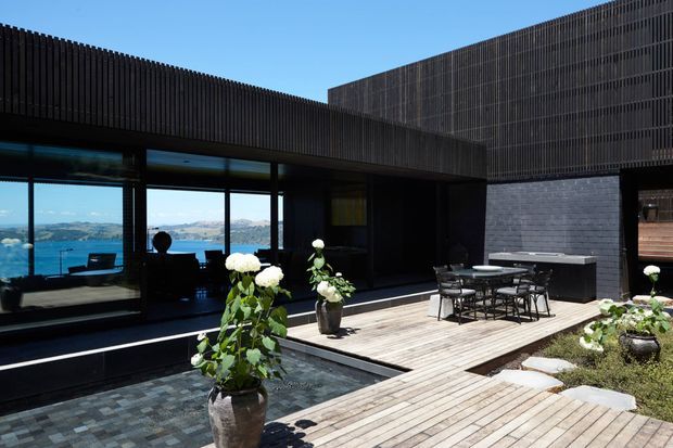 Modern dark-clad residential exterior with a timber-decked courtyard terrace, outdoor dining area, built-in barbecue or counter, reflective water feature, and large glazed living spaces opening toward a blue waterfront landscape. Vertical screening and strong horizontal roof forms create a private, contemporary architectural composition.