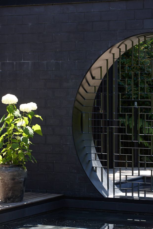 A dramatic architectural exterior detail featuring a dark painted brick wall with a large circular opening secured by a black metal grid. The opening reveals a small paved area and greenery beyond. In the foreground is a shallow dark reflecting pool or water feature, while a large textured planter with white flowering foliage sits on the edge. The composition is contemporary, graphic, and high-contrast, with strong geometric forms and deep charcoal tones.