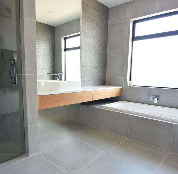 Contemporary bathroom featuring large grey tiled walls and floor, a frameless glass shower, wall-mounted timber vanity with a white vessel basin, freestanding-style bathtub, black-framed windows, and chrome fixtures. The space is bright, clean, and minimal.