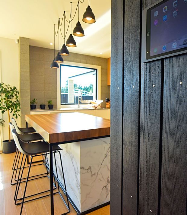 Contemporary kitchen with a large timber island featuring a white marble-look waterfall end, black bar stools, and a row of sculptural black pendant lights. The kitchen includes a dark vertical timber feature wall with an integrated wall-mounted screen, warm timber flooring, pale cabinetry, a stone-look backsplash, and a large window overlooking greenery.