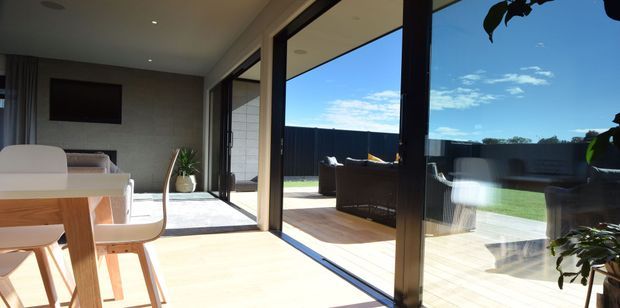 Bright contemporary open-plan dining and living area with a timber dining table and light upholstered chairs in the foreground. Floor-to-ceiling glazed sliding doors open to a timber deck and lawn, with an outdoor seating area visible beyond. The interior has pale timber flooring, a neutral grey feature wall, integrated television, recessed ceiling lighting, and strong indoor-outdoor connection.