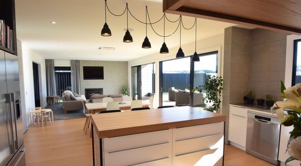 Open-plan contemporary kitchen and living area with a large timber island, white drawer cabinetry, stainless-steel appliances, pendant lighting, and adjoining lounge and dining spaces. Full-height glazing opens to an outdoor area, while a textured stone feature wall adds warmth and definition.