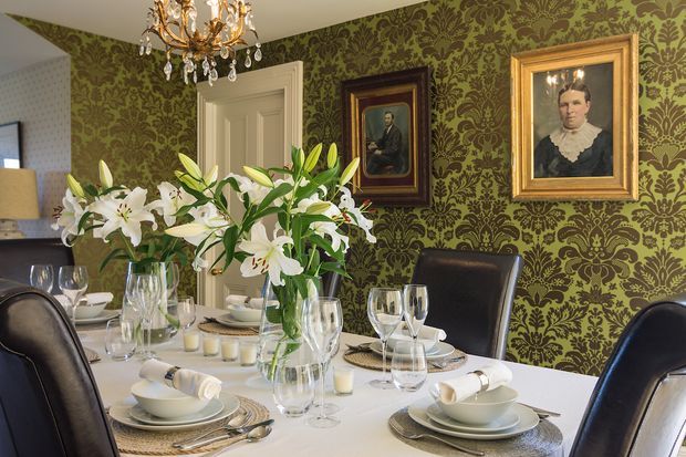 An elegant formal dining room with a fully set white dining table, black leather dining chairs, crystal glassware, layered place settings, and large arrangements of white lilies. The room features ornate olive-green damask wallpaper, framed portrait artwork, a white paneled door, and a decorative crystal chandelier, creating a richly traditional and refined atmosphere.