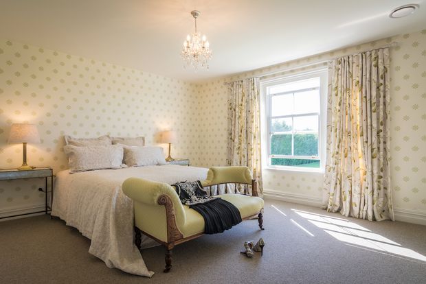 Bright, elegant bedroom with a large upholstered bed, pale green buttoned chaise lounge, bedside lamps, patterned wallpaper, full-length patterned curtains, chandelier, and carpeted floor. A tall window provides abundant natural light and overlooks greenery.