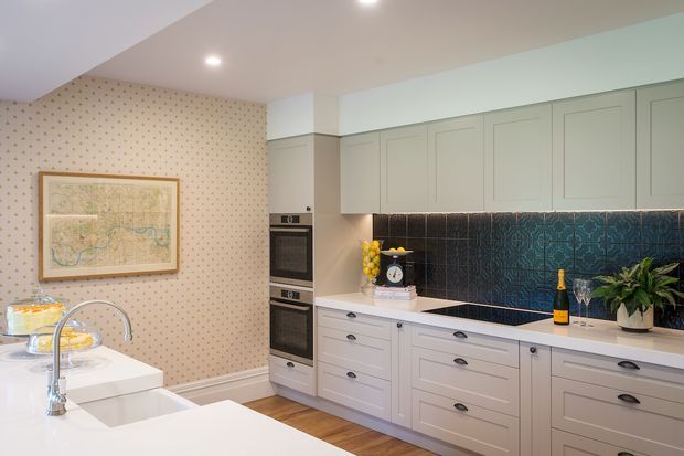 A bright, spacious contemporary kitchen with pale grey-green shaker-style cabinetry, white benchtops, a deep teal patterned tile splashback, integrated double ovens, and timber flooring. A large island with a gooseneck tap and double sink occupies the foreground. Patterned wallpaper, framed artwork, decorative glassware, and indoor plants add character.