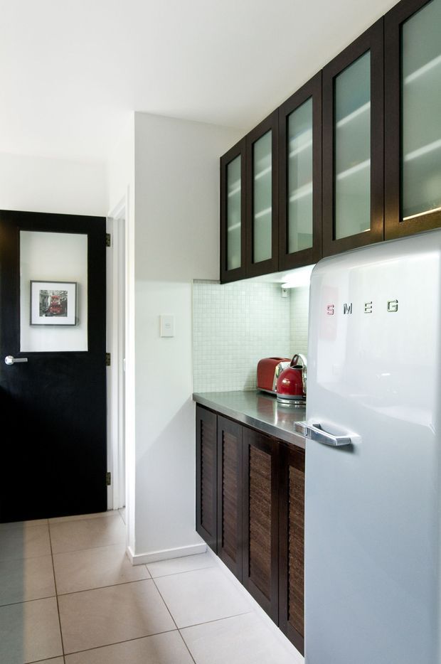 Compact, well-lit kitchen or kitchenette with dark timber cabinetry, glass-front upper cupboards, a stainless-steel benchtop, small square tiled splashback, and a prominent retro white Smeg refrigerator. A black-framed door opens to the adjacent area, and the floor is finished in large light ceramic tiles.
