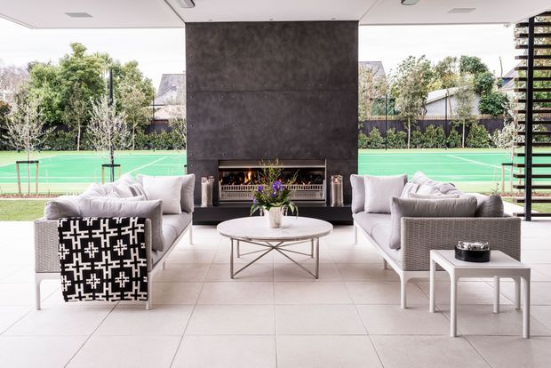 Open-plan covered outdoor living area with pale tiled flooring, a large dark stone or concrete fireplace feature wall, and symmetrical grey upholstered seating arranged around a round coffee table. The space opens directly onto a bright green tennis court and landscaped suburban surroundings, with a contemporary ceiling and dark horizontal privacy screening at the side.