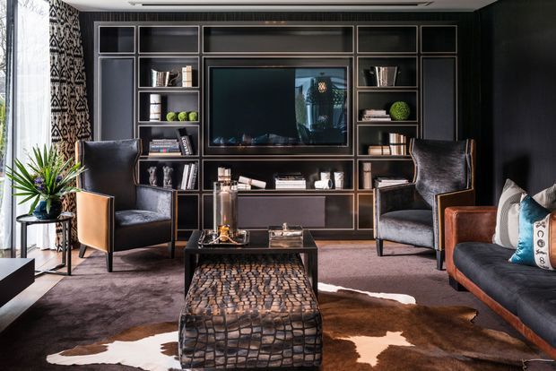 Sophisticated dark-toned media living room with a full-height built-in entertainment wall, central television, open shelving, and integrated cabinetry. Two leather wingback chairs face a low textured coffee table, with a leather sofa, patterned cushions, cowhide rug, and indoor plant. Decorative geometric wall covering and large windows add visual contrast and natural light.