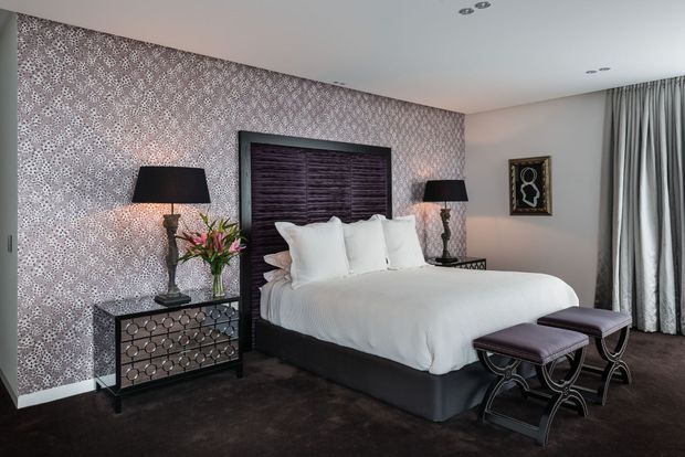 Elegant bedroom with a large upholstered bed, dark wood headboard, patterned feature wallpaper, mirrored geometric bedside cabinet, matching dark bedside lamps, upholstered bench seating, grey curtains, and dark carpet. The room has a refined, luxurious and slightly dramatic character.
