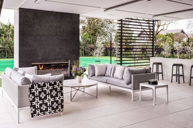 A bright, open-plan covered outdoor lounge with pale tiled flooring, white structural surfaces, grey sofas and armchairs, a dark feature fireplace wall, and a linear outdoor fire. A slatted black screen separates the seating area from a green tennis court and landscaped garden. A compact bar-height counter with stools sits to the right.