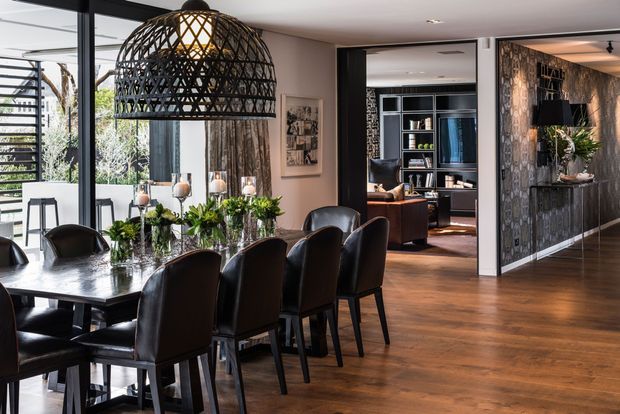A refined open-plan dining area with a long dark dining table surrounded by black upholstered chairs, a large woven pendant light, and a floral table arrangement. Warm timber flooring contrasts with dark feature walls and cabinetry. Full-height glazing opens toward an outdoor area, while a living room and built-in shelving are visible beyond the dining space.
