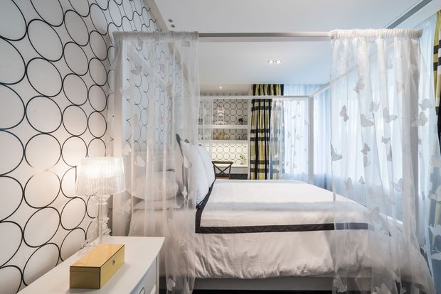 Bright, playful bedroom featuring a four-poster-style bed draped with sheer white curtains, bold oversized black circular wallpaper, patterned textiles, and a white bedside cabinet with a lamp. The room has layered translucent curtains, patterned shelving, and a light contemporary decorative scheme.