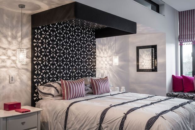 A contemporary bedroom with a low-profile bed dressed in white bedding and striped decorative cushions. A dramatic black geometric patterned feature wall sits behind the bed beneath a large dark canopy-like architectural element. Textured pendant lights cast patterned shadows across pale walls, while a vivid magenta window seat and striped Roman blind add colour. The room has a bold, graphic and refined design.
