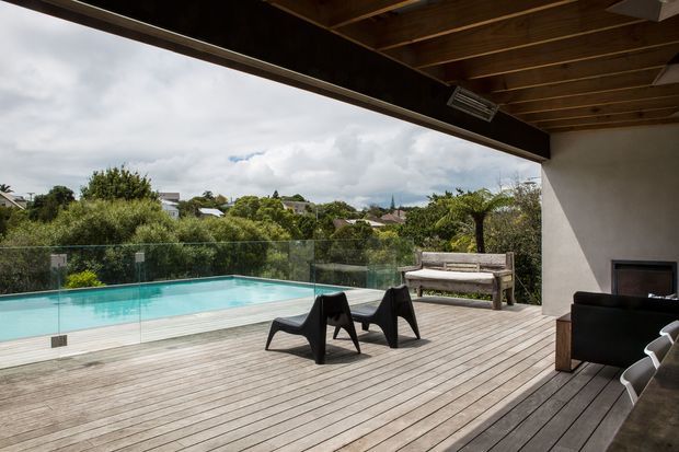 A covered outdoor terrace with a timber-lined ceiling and wide timber decking overlooking a turquoise swimming pool. Frameless glass balustrades separate the deck from the pool, while dark lounge chairs, a bench, and an outdoor fireplace seating area create a contemporary entertaining space. Suburban greenery and houses are visible beyond the pool.
