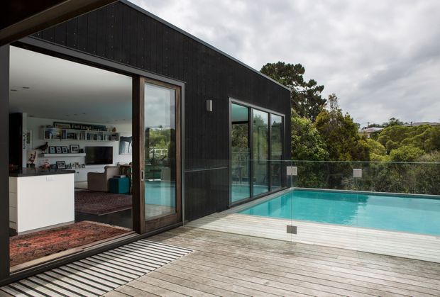 A contemporary dark-clad house with a pitched gable roof, expansive glazed openings, and a timber deck surrounding a turquoise swimming pool. The view looks from the outdoor deck into a bright living room through open sliding doors, with a frameless glass pool balustrade and leafy suburban surroundings.