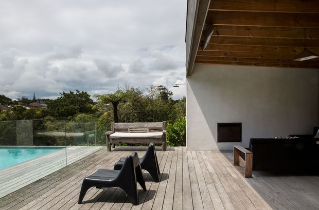 A spacious outdoor timber deck beside a turquoise swimming pool, with frameless glass pool fencing, black outdoor lounge chairs, a timber bench, and a covered white-rendered pavilion. The pavilion has a warm timber-lined ceiling, exposed roof structure, and a built-in dark fireplace or firebox. Lush vegetation and neighbouring rooftops are visible beyond the deck.