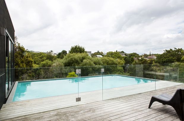 Contemporary elevated outdoor terrace with a rectangular swimming pool, pale timber decking, and frameless glass balustrade. The view looks across dense suburban greenery toward neighbouring homes under an overcast sky; a dark modern building edge and outdoor lounge chair frame the scene.