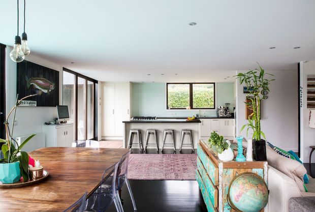 Open-plan residential interior showing a combined dining, kitchen and living area. A large timber dining table occupies the foreground, with a compact kitchen and breakfast island with four stools at the rear. The room features dark timber flooring, white cabinetry, extensive black-framed glazing, pendant lights, indoor plants, and a colourful distressed timber console separating the dining and lounge zones.