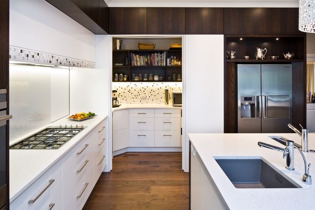 Contemporary kitchen with white flat-panel cabinetry, a large island with integrated sink, stainless-steel appliances, dark timber cabinetry and shelving, warm timber flooring, and mosaic tile splashbacks. A walk-in pantry is visible at the rear.