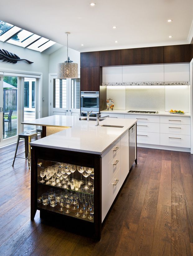 Bright contemporary kitchen with a large white island, integrated sink and appliance cabinetry, dark timber overhead joinery, white drawers, and a mosaic tile splashback. The island includes open glassware storage, while skylights, recessed lighting, and glazed doors provide abundant natural light. Dark timber flooring and a decorative pendant add warmth and contrast.