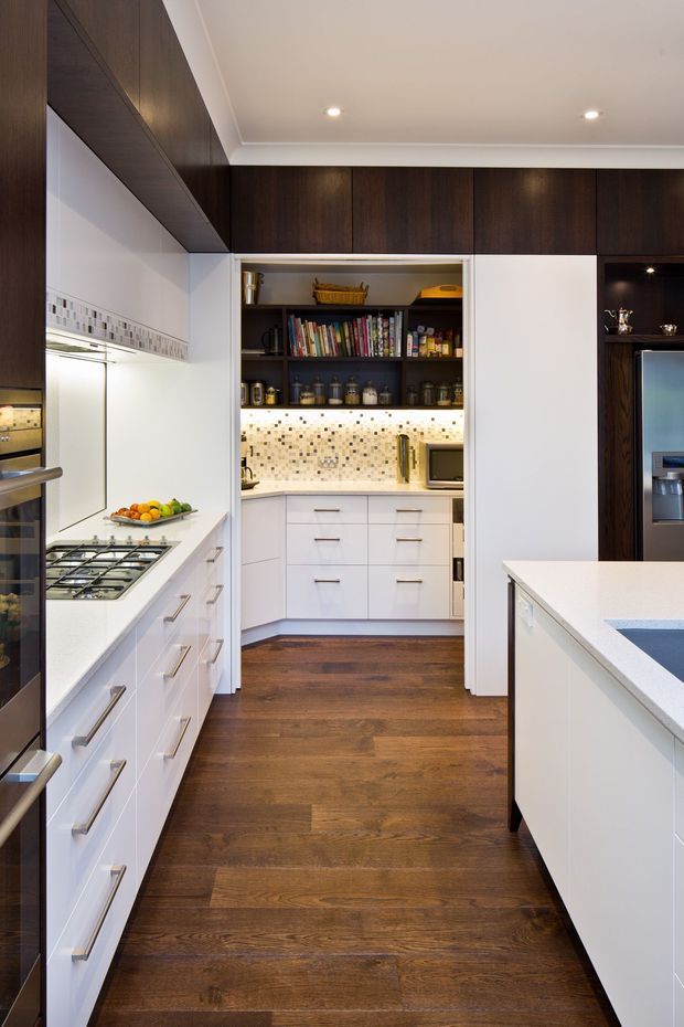 A contemporary galley-style kitchen with white lower cabinetry, dark timber overhead cabinetry and wall panels, white benchtops, stainless steel appliances, and a mosaic tile splashback. The room features a central white island, integrated storage, recessed ceiling lighting, and dark timber flooring.
