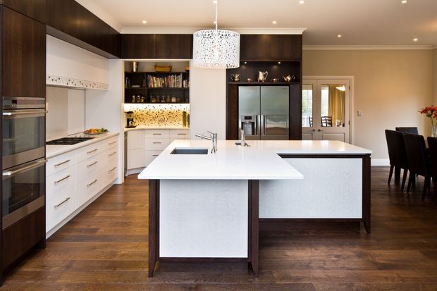 A spacious open-plan kitchen and dining area with two large white waterfall-edge islands, dark timber cabinetry, integrated stainless-steel appliances, a decorative tiled splashback, and warm timber flooring. A dining table and chairs are visible to the right, with a pendant light centred over the island.