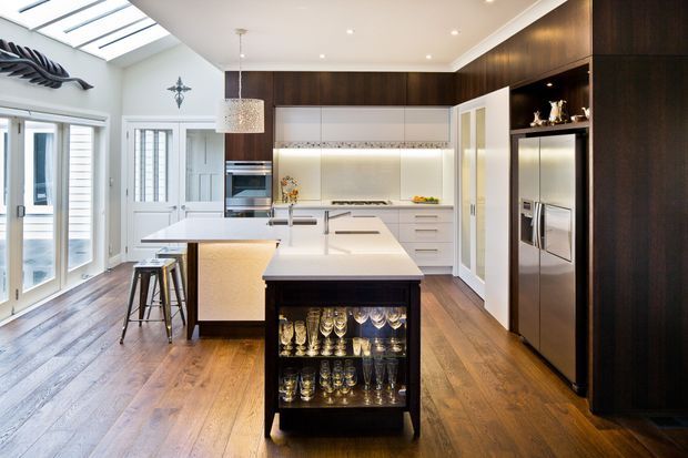 Bright, spacious contemporary kitchen with a large white island, integrated illuminated display storage, dark timber cabinetry, stainless-steel appliances, white cabinetry, timber flooring, and abundant natural light from skylights and glazed doors. A smaller island or bar area with stools sits behind the main island.