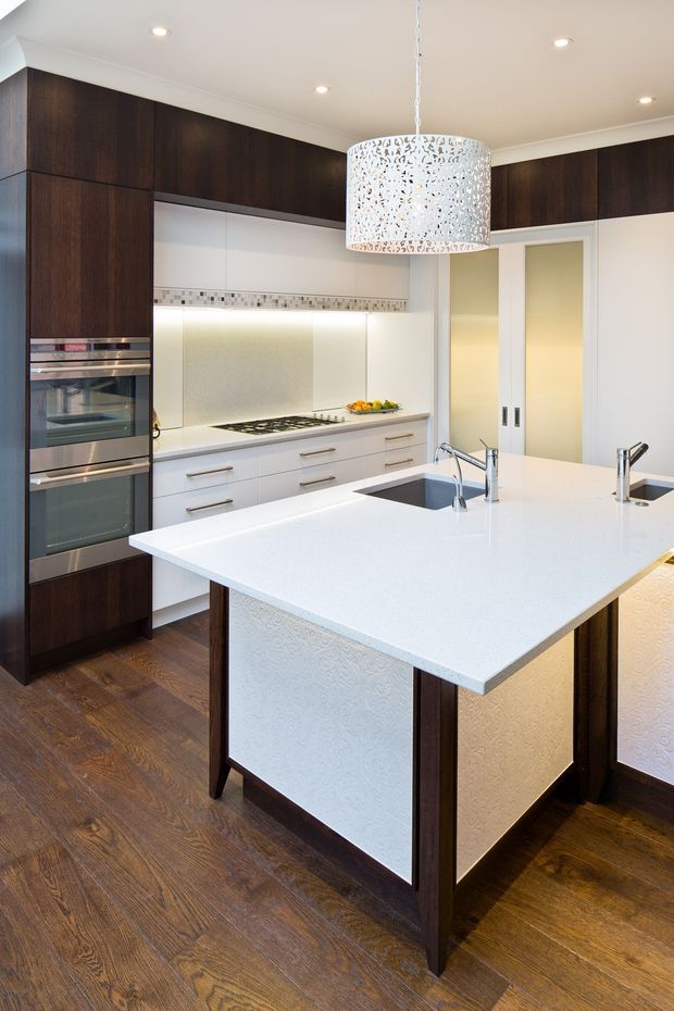 A bright, contemporary kitchen featuring a large white island with integrated sink and cooktop, dark timber cabinetry, white lower cabinets, stainless-steel appliances, patterned backsplash detailing, and a decorative pendant light. Warm timber flooring contrasts with the white benchtops and cabinetry.