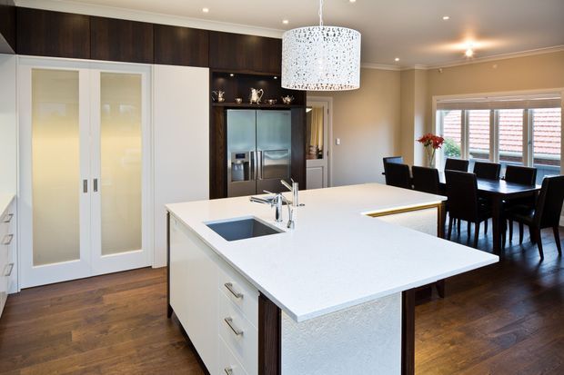 Bright, spacious open-plan kitchen and dining area with a large white island, integrated sink and cabinetry, dark timber overhead and appliance joinery, warm timber flooring, and a white decorative pendant light. A dining table sits beside large windows with horizontal blinds, while glass-panelled doors and built-in storage line the opposite wall.