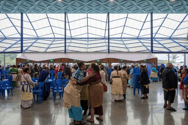 A large, brightly lit communal interior with a dramatic geometric glazed roof supported by blue steel framing. A patterned blue ceiling feature spans the foreground, while numerous people gather around rows of blue plastic chairs and a long timber-clad service or seating structure. The polished concrete floor and expansive open layout suggest a public hall, church, community venue, or event space.