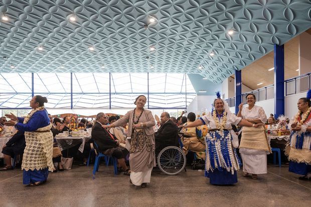 A spacious public interior filled with seated guests and performers in traditional Pacific Island attire. The room features a highly distinctive sculptural geometric patterned ceiling, polished concrete flooring, extensive glazed walls or roof glazing, and blue structural columns. The image appears to capture a cultural or ceremonial event in a large commercial or community venue.