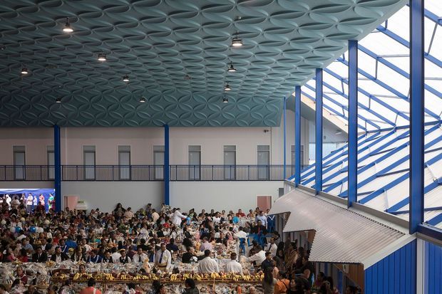 A large, brightly lit commercial atrium or event hall filled with a seated crowd and banquet tables. The space features a dramatic repeating oval-pattern ceiling, exposed blue structural framing, extensive glazed walls and roof-like clerestory glazing, upper-level balcony railings, and a large sloped metal-clad architectural element along the right side.