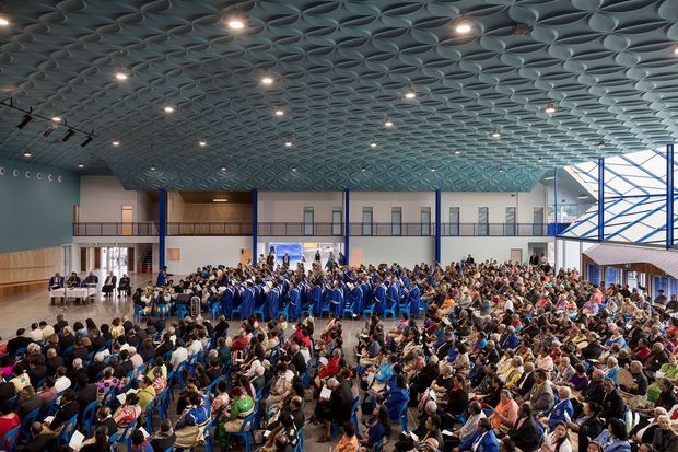 A large contemporary multi-purpose hall or auditorium filled with a seated audience and a graduation ceremony in progress. The space features a dramatic vaulted ceiling covered in repeating geometric acoustic panels, numerous recessed and track lights, blue structural columns and balustrades, timber wall cladding, and a two-level gallery around the perimeter. Extensive glazing and a bright glazed stair or circulation volume are visible on the right.