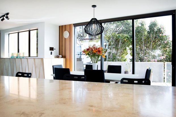 Bright contemporary open-plan dining and kitchen area viewed across a large pale stone island or benchtop. The room features black-framed floor-to-ceiling glazing, white walls and ceiling, vertical timber wall panelling, black dining chairs, and a sculptural black wire pendant light. A garden is visible beyond the glazing.