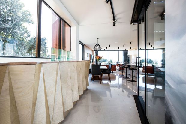 Bright, open-plan contemporary interior with a long sculptural light-wood island or counter in the foreground, polished pale flooring, extensive floor-to-ceiling glazing, and black-framed glass partitions. A dining and lounge area with dark furniture sits beyond the counter, while linear track lighting and a geometric pendant add a modern graphic element.