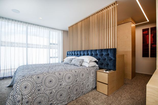 A contemporary bedroom featuring a blue button-tufted upholstered bedhead integrated with timber bedside joinery and a vertical timber-slat room divider. The room has floor-to-ceiling sheer glazing with curtains, pale timber built-ins, patterned monochrome bedding, carpeted flooring, and recessed linear ceiling lighting.