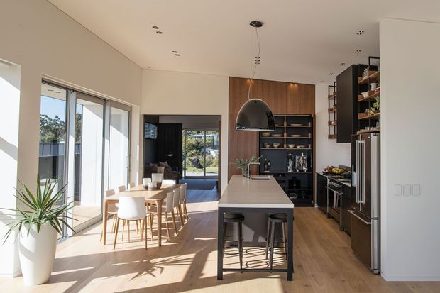 Bright open-plan kitchen and dining area with a large central island, white stone benchtop, dark timber cabinetry, integrated shelving, stainless-steel appliances, and a dining table beside expansive glazed doors. The space has warm timber flooring, a sculptural dark pendant light, and a clean contemporary fit-out.