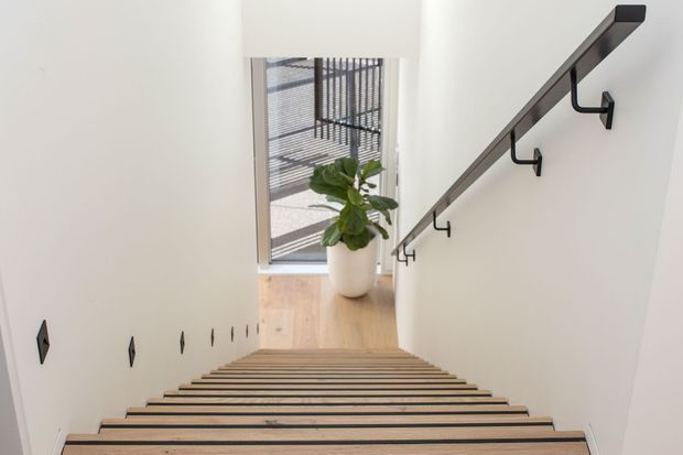 View down a modern stairway with light timber treads, white walls, dark metal edge detailing and a dark timber or metal handrail with integrated hooks. A tall window brings in daylight, with a potted plant on the landing below.