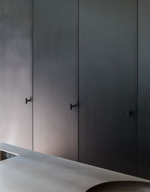 Close-up of a dark, minimalist built-in cabinetry wall with tall flush doors and small black pull handles. A pale grey stone benchtop with a sculptural curved edge occupies the foreground, with part of a stainless-steel sink visible at the left. The image is tightly framed and moody, emphasizing the cabinetry and benchtop rather than the wider room.