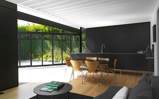 Contemporary open-plan kitchen and dining area with extensive black built-in cabinetry, a long island, and a dining table with timber-toned chairs. Full-height black-framed glazing opens to a leafy garden, while a white exposed-beam ceiling contrasts with warm timber flooring. A portion of a dark sofa and round coffee table is visible in the foreground.