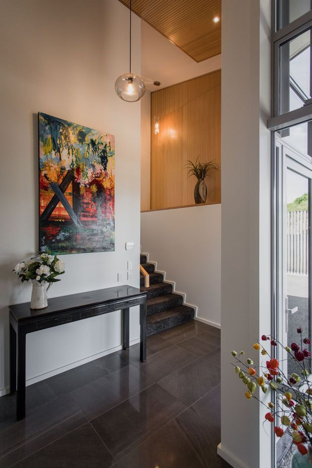 A refined residential entryway with a tall ceiling and stairwell. The space features dark tiled flooring, a slim black console table, a colourful abstract artwork, a sculptural pendant light, and a staircase with dark carpeted treads. Warm vertical timber panelling continues up the stair landing and across the ceiling, while large framed glazing brings in daylight and provides an exterior view.