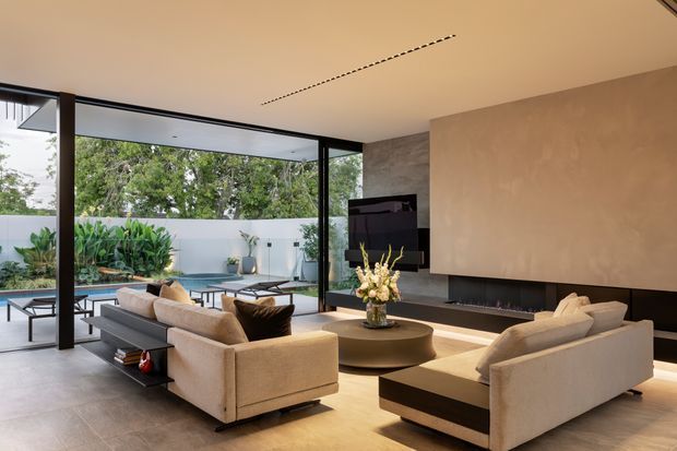 A sophisticated contemporary living room with floor-to-ceiling black-framed glazing opening onto a planted courtyard and pool area. The room features a large textured stone or plaster feature wall, a wide linear fireplace beneath a wall-mounted television, low neutral-toned sofas, a round coffee table, and integrated dark cabinetry. Warm indirect lighting and restrained natural materials create a refined indoor-outdoor atmosphere.